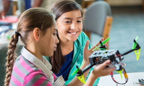 Elementary age Hispanic little girls are using drones during after school science club or program. Students are studying science, technology, engineering, and math in public elementary school library makerspace.
